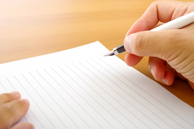 Person writing with a white fountain pen on ruled notebook paper at a wooden desk surface.