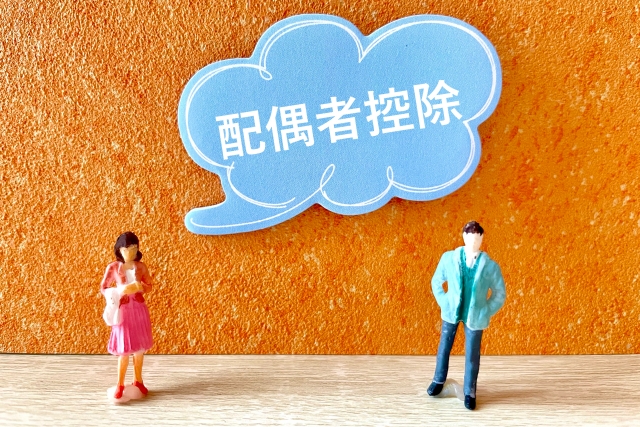 Miniature man and woman stand beside a blue cloud-shaped sign with 配偶者控除, representing a government spouse deduction policy against an orange wall.