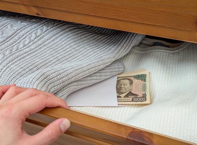 Hand pulling a white envelope from under a blue knit blanket in a wooden drawer; a 10,000 yen banknote peeks out with portrait visible.