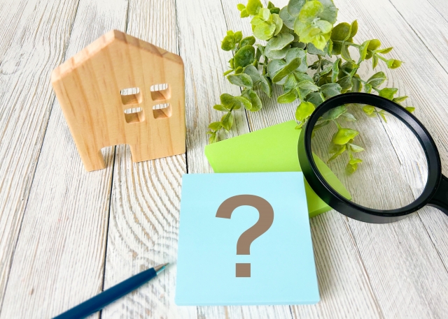Blue sticky note with a question mark beside a wooden house model, magnifying glass, plant, and pencil on a light wood surface.