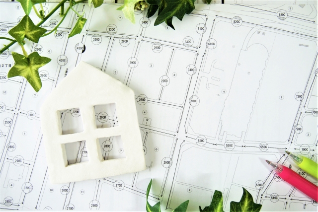 White miniature house on architectural blueprints, surrounded by green ivy and a pink pencil, symbolizing home design planning.